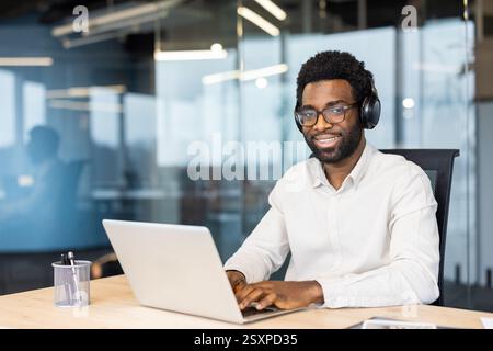 Young man wearing headphones, glasses, and a white shirt works on his laptop in an office setting, smiling at the camera. Stock Photo