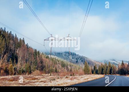 High-voltage electric transmission tower surrounded with forest in mountains. Electricity pylon. Winter landscape of power lines by road Stock Photo