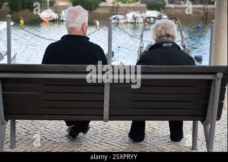 Portugal, Algarve; Fuseta. Elderly couple on a bench looking at boats on the water. Stock Photo