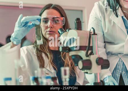 Young female scientist analyzing a sample in a laboratory. Wearing safety glasses and gloves, she works with a microscope, representing biotechnology, Stock Photo