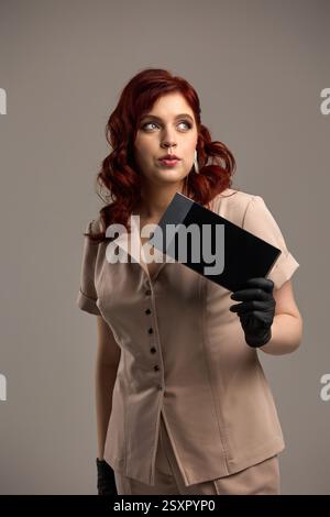 Red-haired woman holding black envelope, dressed in beige attire and gloves, gazing to the side, confident style concept Stock Photo