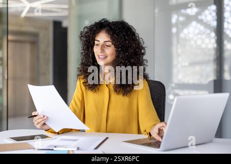 Successful and joyful woman at workplace checking financial reports and contracts. Businesswoman doing paperwork inside office, using laptop. Financie Stock Photo