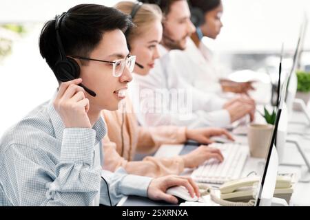 Asian helpdesk representative in headphones with his colleagues providing tech support services to customers, copy space Stock Photo