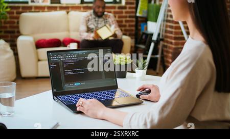 Asian female programmer works on coding software in a home office setup, typing code on a laptop. IT expert uses digital tools for multitasking, software development and app development. Camera A. Stock Photo