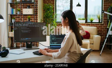 Asian programmer seen typing code for a user interface design on dual monitors, working in a digital workspace. Woman coder working on software engineering and front end development. Camera A. Stock Photo