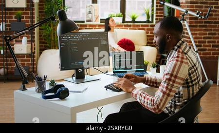 Young man seen programming software code on his laptop in a sleek home office. Workspace with dual monitors helps with software engineering, web development and coding productivity. Camera A. Stock Photo