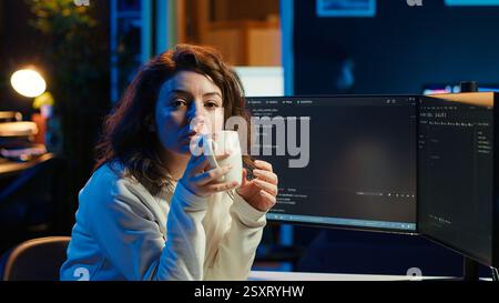 Portrait of smiling IT specialist drinking coffee while looking for company security vulnerabilities and preventing malware infections. Programmer enjoying hot beverage while writing code, camera A Stock Photo