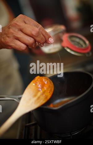Cooking, salt and pot with hand of chef in kitchen of home to flavor or season food closeup. Ingredients, recipe and wooden spoon with fingers of Stock Photo