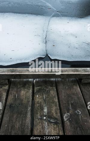 On a quiet winter morning, a wooden pier extends over a lake with a stark division of cracked ice and open water. The scene captures the peacefulness Stock Photo
