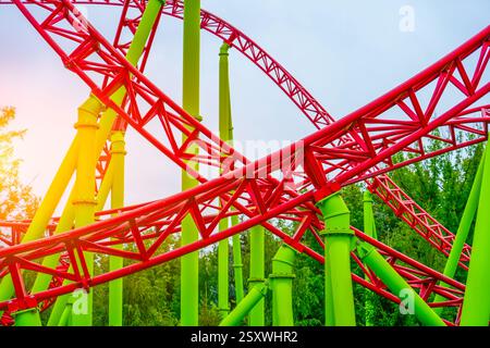 Vibrant red and green roller coaster tracks twist and turn against a cloudy sky. The structure creates a thrilling visual surrounded by lush greenery Stock Photo