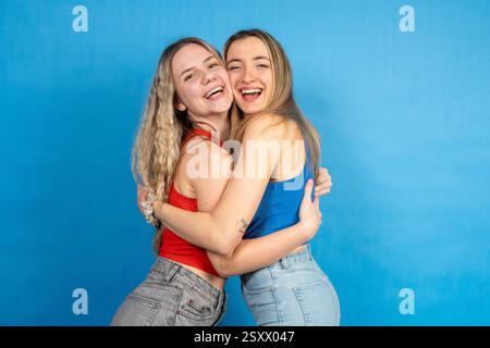 Two cheerful female friends are hugging and laughing, expressing their joy and connection on a vibrant blue backdrop Stock Photo
