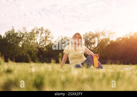Beautiful background. Sitting with bottle. Senior woman having nice weekend outdoors on the field at sunny day. Stock Photo