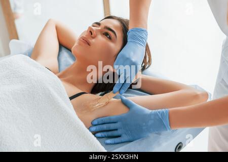 Putting cream on the armpits. Woman is having sugaring procedure in the clinic by female doctor. Stock Photo