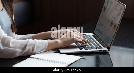 Professional Asian woman analyzing data on a laptop while taking notes in a modern office. Stock Photo