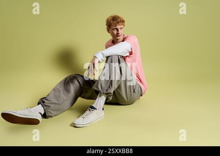 A fashionable young man with curly hair relaxes in a stylish outfit on a smooth pastel surface. Stock Photo