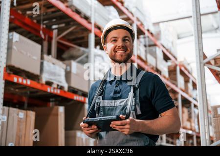 With digital tablet in hands. Employee in uniform is working in the storage at daytime. Stock Photo
