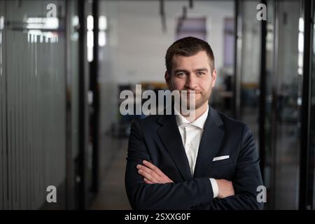 Confident young businessman, manager, or entrepreneur in a modern office, wearing a suit with arms crossed and a friendly smile. Leadership, corporate Stock Photo