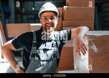 Leaning on the box and smiling. Employee in uniform is working in the storage at daytime. Stock Photo
