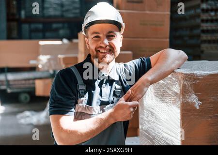 Leaning on the box and smiling. Employee in uniform is working in the storage at daytime. Stock Photo