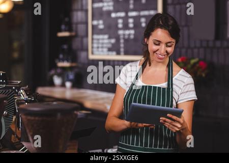 Barista in striped apron using tablet in cozy coffee shop, smiling warmly Stock Photo