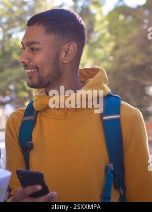 Smiling young man in yellow hoodie holding smartphone outdoors with backpack Stock Photo