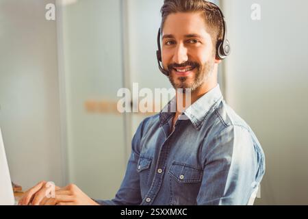 Smiling man in denim shirt using headset while working at office desk Stock Photo