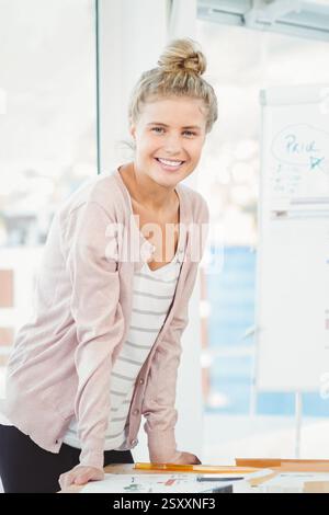 Smiling woman in office leaning on desk with charts, looking confident Stock Photo