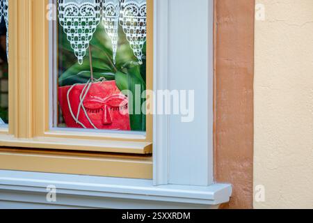 A red flower vaste stands on the windowsill behind a window. It is shaped like a human face, with a nose, mouth and two closed eyes with eyelashes. Stock Photo