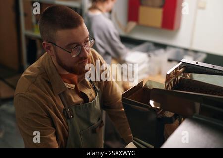 High angle shot of young male worker in apron glancing at coffee grinder while his colleague zipping packs of ground coffee Stock Photo