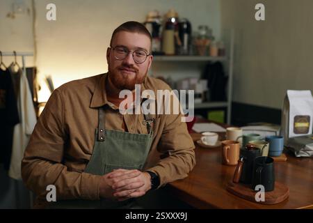 Medium close up portrait of Caucasian young adult entrepreneur smiling while sitting at table with handmade cups for coffee Stock Photo