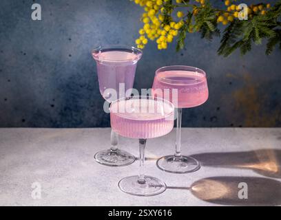 Three pink cocktails in elegant glasses stand on a table, accompanied by mimosa flowers, creating a festive atmosphere Stock Photo