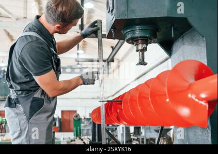 Measuring red colored sharp part. Man in uniform is in workstation developing details of agriculture technique. Stock Photo