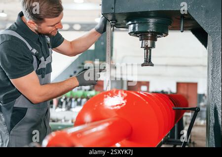 Measuring red colored sharp part. Man in uniform is in workstation developing details of agriculture technique. Stock Photo
