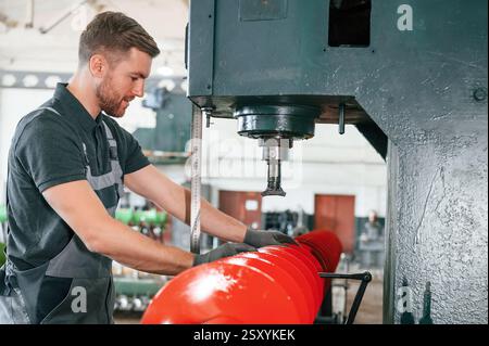 Measuring red colored sharp part. Man in uniform is in workstation developing details of agriculture technique. Stock Photo