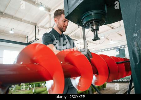 Measuring red colored sharp part. Man in uniform is in workstation developing details of agriculture technique. Stock Photo