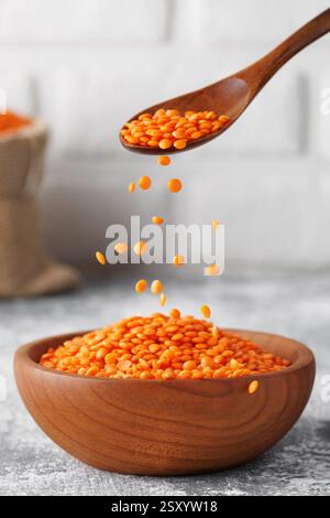 Red lentils pouring from wooden spoon into rustic bowl standing on grey kitchen table against white brick wall Stock Photo