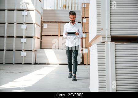 Holding tablet and checking the data. Storage worker is in the warehouse with bunch of products. Stock Photo