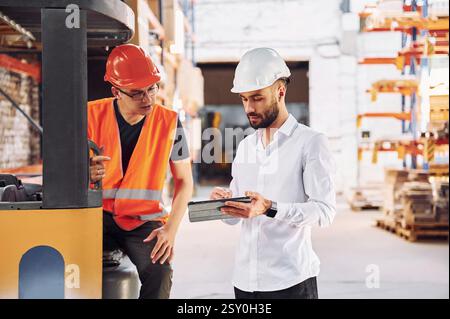 With tablet and cargo moving transport. Two storage workers is in the warehouse. Stock Photo