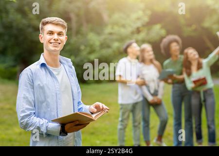 Education First. Student Guy Posing With Book While His Classmates Taking Selfie Stock Photo
