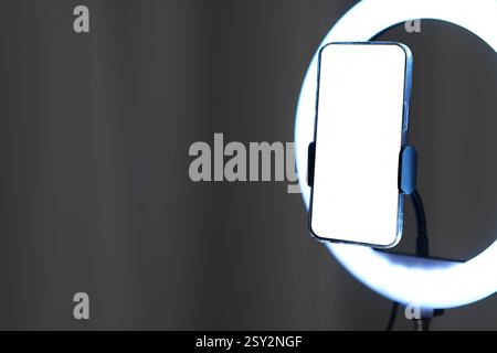 Glowing ring lamp with smartphone near grey curtain, closeup. Space for text Stock Photo
