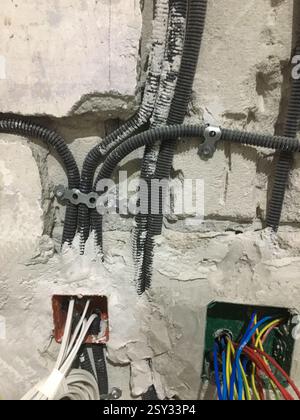 A wall under construction features gray corrugated conduits, metal brackets, and open electrical boxes with exposed wires. Plaster and concrete are vi Stock Photo
