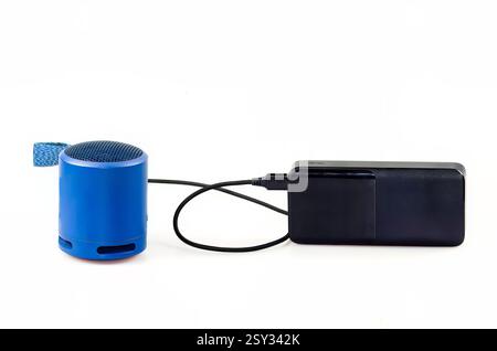 Compact blue cylindrical speaker with mesh top and fabric loop connected to a sleek black power bank via cable, set against a white background. Stock Photo