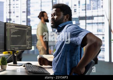 Worker stretching to relieve muscle strain while conducting code review to fix bugs, identifying technical issues with software. Man in office suffering back pain from bad posture, optimizing code Stock Photo