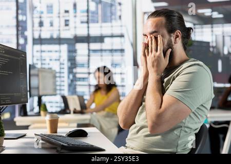 Exhausted man at work rubbing eyes, creating machine learning models that can process and analyze data. Tired IT expert programming in office, struggling to stay awake after pulling all nighter Stock Photo
