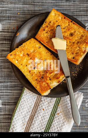 Two toasted bread slices with a table knife holding a piece of butter, placed on a dark plate with a white cotton napkin beside it. Stock Photo