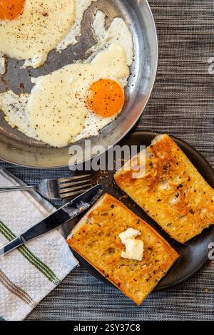 Two toasted bread slices with  butter on a dark plate, accompanied by a knife and fork on a napkin, with a pan of fried eggs sprinkled with black pepp Stock Photo