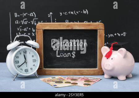 Chalkboard with text EDUCATION LOANS, piggy bank, alarm clock and money on table near blackboard Stock Photo