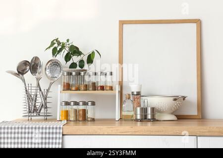 Shelf with different spices, plant, blank frame and utensils on counter near light wall in kitchen Stock Photo