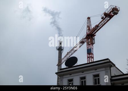 A low-angle shot of a red construction crane juxtaposed against a smokestack emitting smoke, set against an overcast sky. Stock Photo