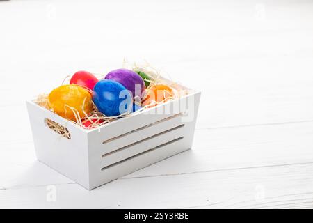 Colored Easter eggs lie in a white wooden box in straw on a white wooden background Stock Photo